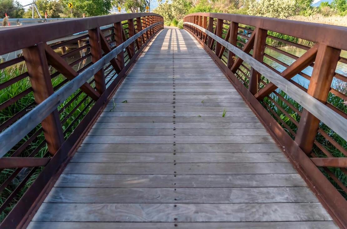 Bridge with wood deck and rusty metal railing over a lake with grassy shore. Trees and road signs can also be seen from the structure on this sunny day.